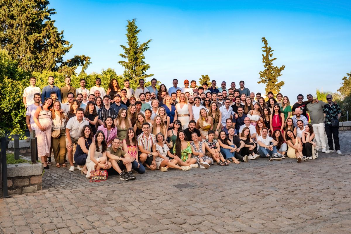 Foto donde hay algo más de 100 personas de pie posando para la foto, fondo con árboles y suelo adoquinado