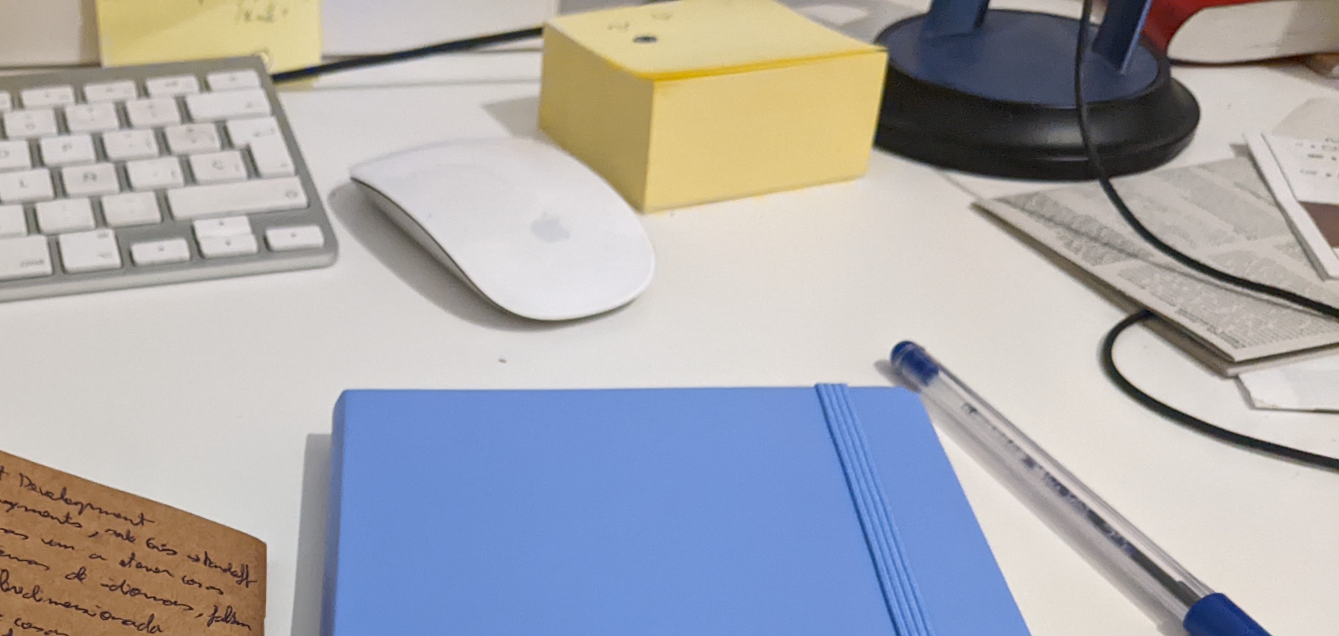 Foto de un escritorio donde hay un teclado, un ratón, un taco de post its, un par de cuadernos pequeños y un boli
